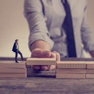 Man solving problems by building bridge with wooden block to span a gap for little businessman walking across in a conceptual retro toned image.